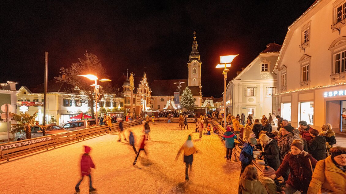 Eine lebhafte Eisbahn voller Schlittschuhläufer unter dem nächtlichen Himmel. Im Hintergrund sind festlich beleuchtete Gebäude und ein Kirchturm zu sehen. | © Rene Strasser