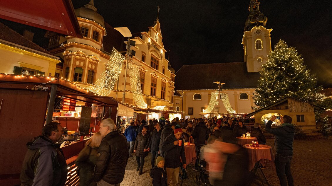 Ein festlicher Weihnachtsmarkt mit zahlreichen Besuchern und schön beleuchteten Ständen. Im Hintergrund sind historische Gebäude und ein geschmückter Baum zu sehen. | © Rene Strasser