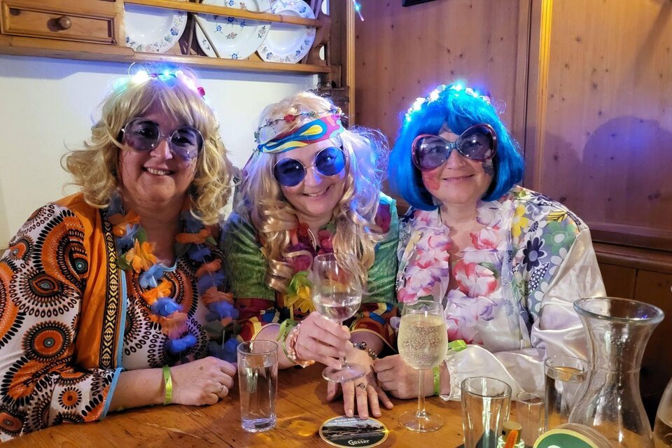 Drei Frauen in bunten Kostümen und Sonnenbrillen sitzen an einem Tisch und lächeln. Sie tragen Blumenketten und halten Gläser in der Hand. | ©  Petra Kirchschlager