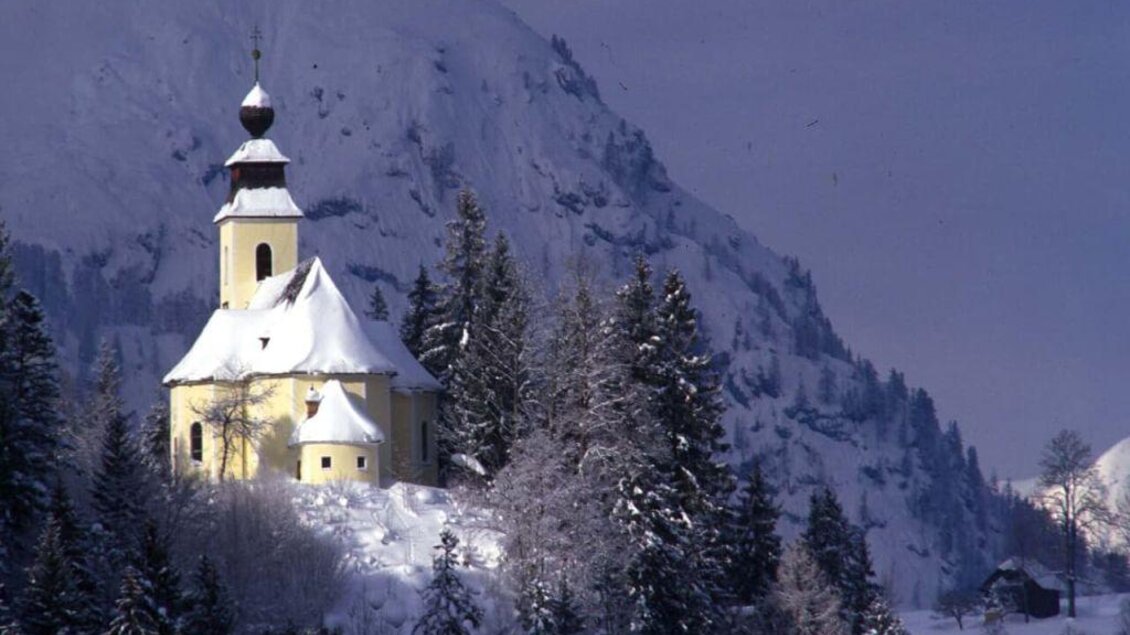 Eine malerische Kirche auf einem verschneiten Hügel, umgeben von frostigen Bäumen. Im Hintergrund erheben sich beeindruckende Berge unter einem bedeckten Himmel. | © TVB Ausseerland - Salzkammergut_Pirker