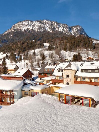 Ein malerisches Bergdorf im Winter mit schneebedeckten Häusern. Im Hintergrund erheben sich majestätische Berge unter einem klaren blauen Himmel. | © Hotel Der Hechl