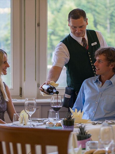 Ein elegantes Restaurant mit einem Kellner, der Wein einschenkt. Ein Paar geniert sich an einem schön gedeckten Tisch mit Blick auf die Natur. | © Florian Loitzl