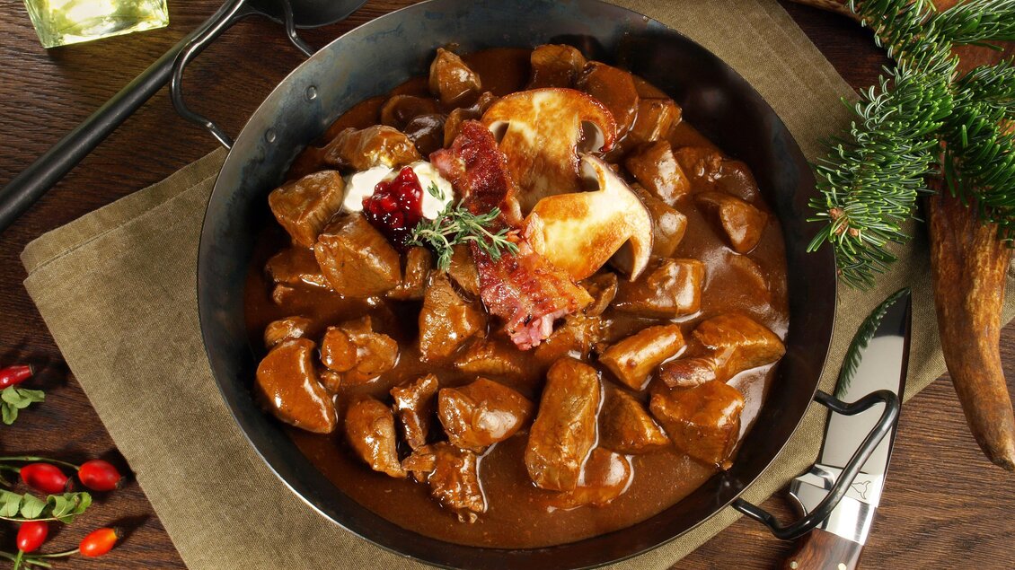 Ein herzhaftes Gulaschgericht in einer Pfanne, garniert mit Blutwurst und frischen Kräutern. Die Speise ist umgeben von rustikalen Tischaccessoires. | © AdobeStock ExQuisine