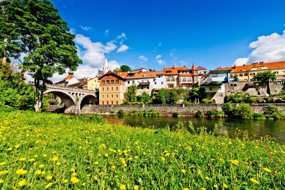 Eine malerische Stadtansicht mit bunten Häusern und einer Brücke über einen Fluss. Die Wiese im Vordergrund ist mit gelben Blumen bedeckt und der Himmel ist klar und blau.