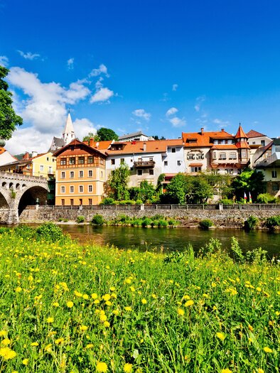 A picturesque town view with colorful houses and a bridge over a river. The meadow in the foreground is covered with yellow flowers and the sky is clear and blue.