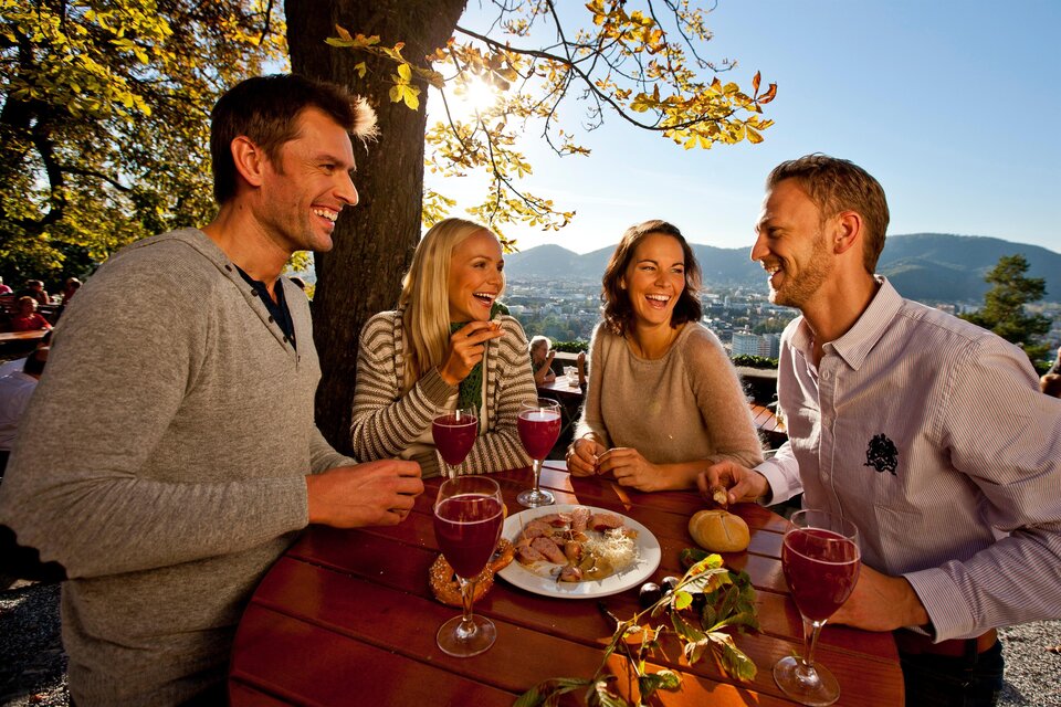 Vier Freunde genießen ein fröhliches Treffen im Freien, umgeben von Bäumen und einem schönen Ausblick. Auf dem Tisch stehen Getränke und Snacks. | © (c) Graz Tourismus - Tom Lamm
