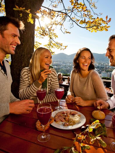 Four friends enjoy a cheerful gathering outdoors, surrounded by trees and a beautiful view. Drinks and snacks are on the table. | © (c) Graz Tourismus - Tom Lamm