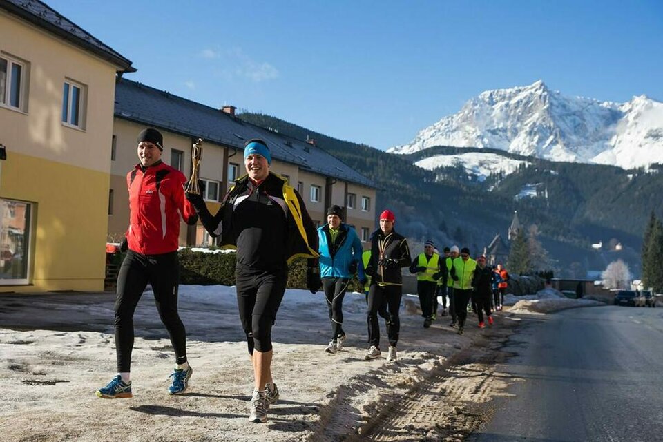 Eine Gruppe von Läufern joggt an einer verschneiten Straße entlang. Im Hintergrund sind Berge und blauer Himmel zu sehen. | © Walter Eisner
