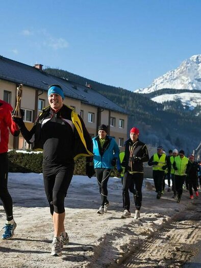 A group of runners jogs along a snow-covered road. In the background, there are mountains and a blue sky. | © Walter Eisner