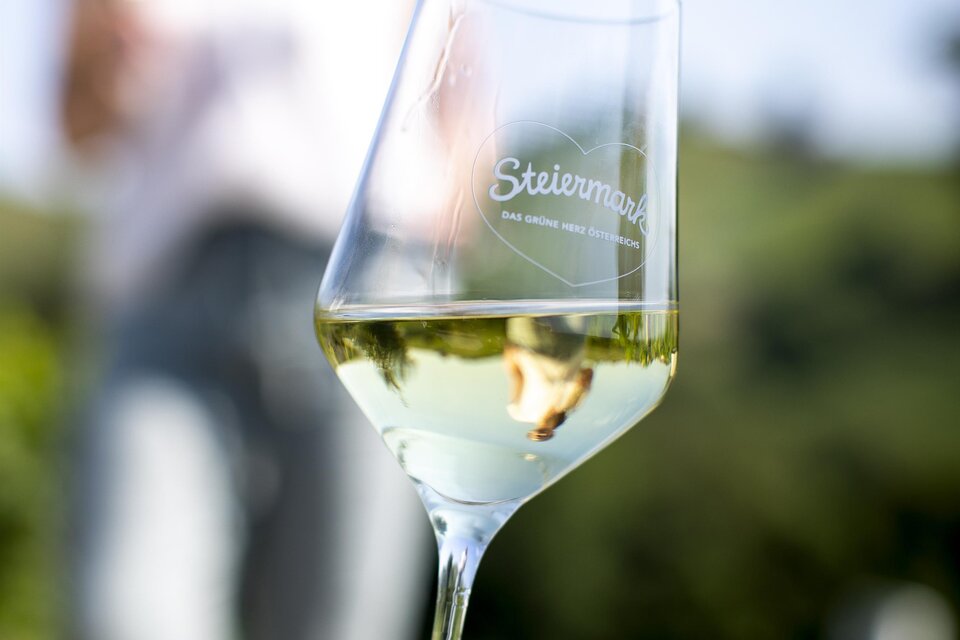 Ein Glas Wein in der Hand, im Hintergrund eine verschwommene Person. Die Szenerie vermittelt eine entspannte, sonnige Atmosphäre im Freien. | © TVB Südsteiermark