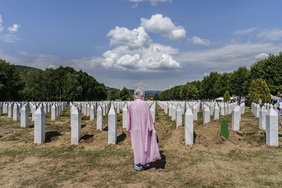 Eine Person in einem rosa Gewand steht inmitten eines Friedhofs mit weißen Grabsteinen. Im Hintergrund sind grüne Bäume und ein blauer Himmel mit Wolken sichtbar. | © Pavelhaus