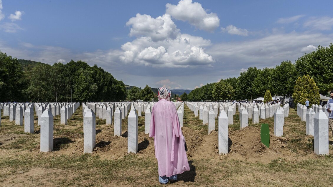 Eine Person in einem rosa Gewand steht inmitten eines Friedhofs mit weißen Grabsteinen. Im Hintergrund sind grüne Bäume und ein blauer Himmel mit Wolken sichtbar. | © Pavelhaus