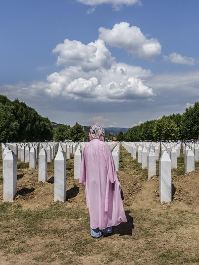 A person in a pink robe stands in the middle of a cemetery with white gravestones. In the background, green trees and a blue sky with clouds can be seen. | © Pavelhaus