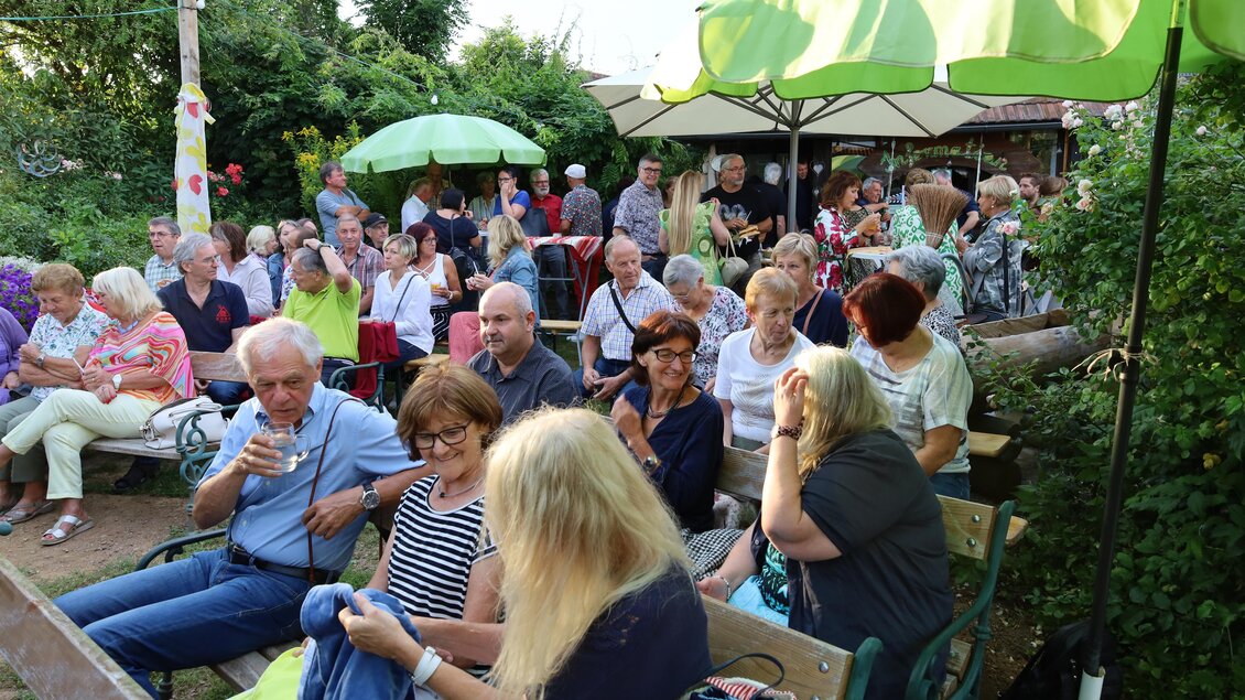 Eine belebte Gartenveranstaltung mit vielen Menschen, die auf Bänken sitzen. Im Hintergrund sind grüne Sonnenschirme und reichlich Vegetation zu sehen. | © Erich Meister