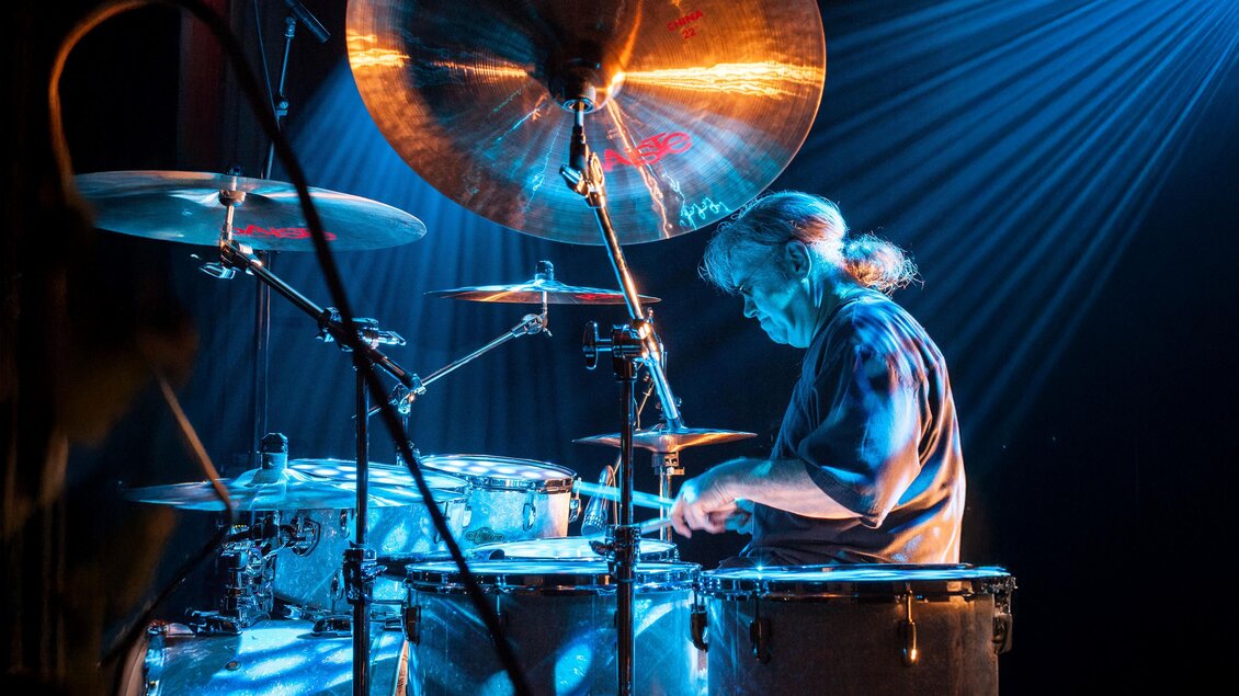 Schlagzeuger spielt auf der Bühne am Drumkit, in blauem Licht und mit sichtbaren Lichtstrahlen; große Becken dominieren den Vordergrund. | © Direkt Bookin Berlin