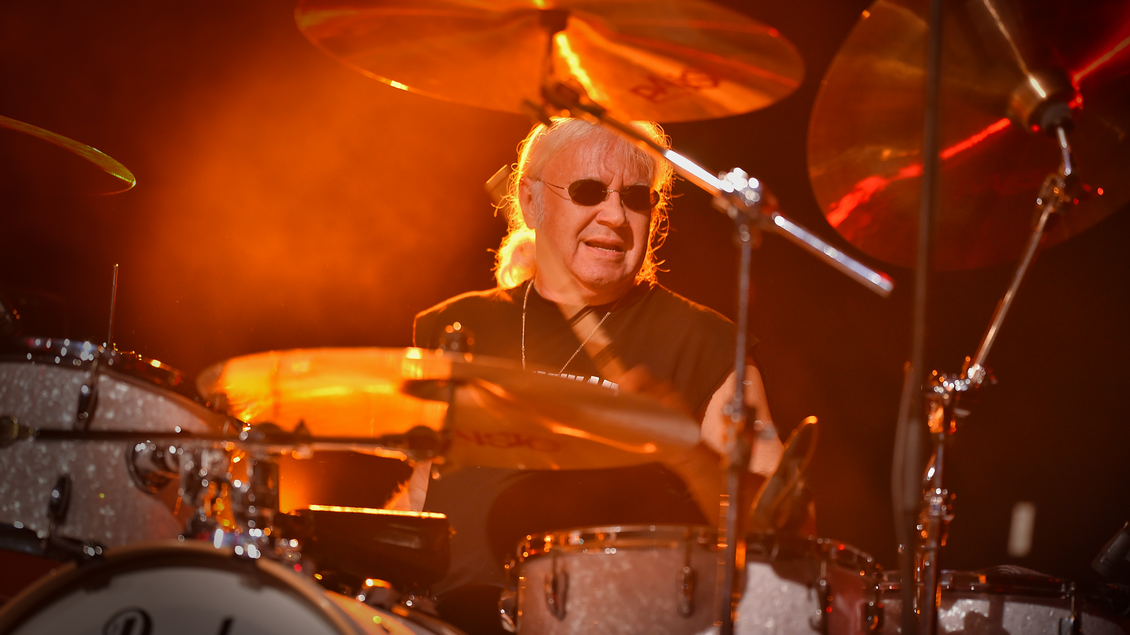 Schlagzeuger am Drumset in warmem, orangefarbenem Bühnenlicht und Nebel; mehrere Becken und Trommeln im Bild, dynamische Konzertstimmung. | © Direkt Booking Berlin