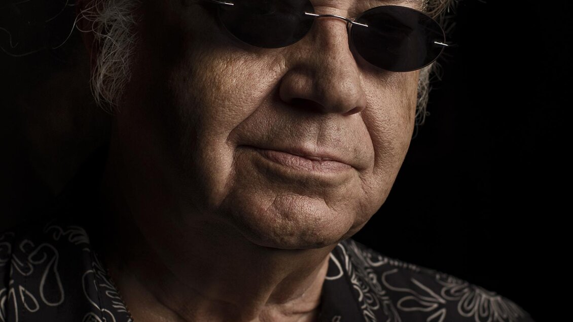 Nahportrait eines Mannes mit hellen Haaren und dunkler Sonnenbrille vor schwarzem Hintergrund; trägt ein gemustertes Hemd, neutraler Gesichtsausdruck. | © Direkt Bookin Berlin