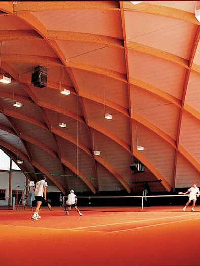 Eine moderne Tennishalle mit einem hohen, gewölbten Dach. Auf dem leuchtend orangefarbenen Platz spielen mehrere Personen Tennis. | © Lederer