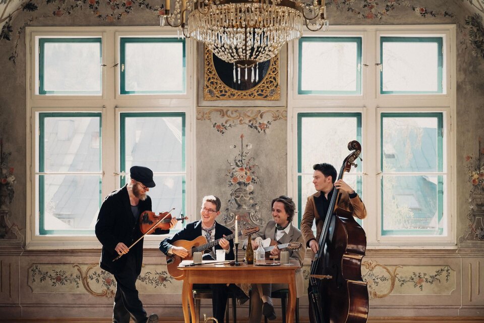 Eine Gruppe von vier Musikern spielt in einem eleganten Raum mit einem Kronleuchter. Der Tisch ist mit Getränken und Instrumenten dekoriert. | © Karl Steinegger