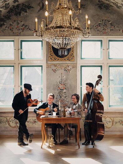 A group of four musicians is playing in an elegant room with a chandelier. The table is decorated with drinks and instruments. | © Karl Steinegger