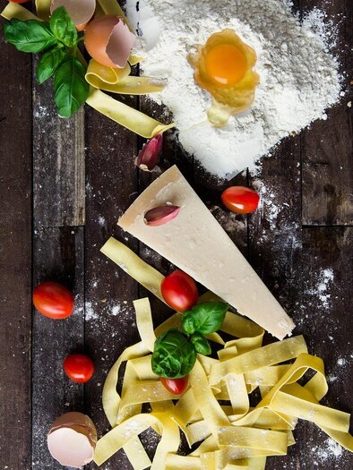 A little pasta, eggs, flour, and fresh ingredients like cherry tomatoes and basil are laid out on a wooden table. The image conveys an inviting atmosphere for Italian cuisine. | © Pixabay
