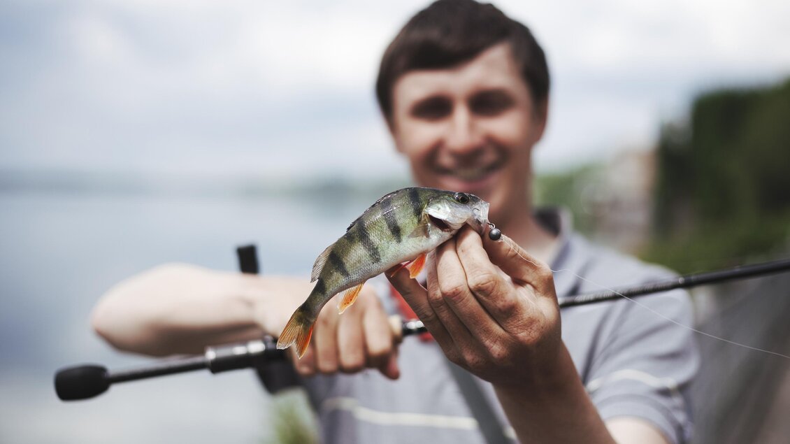 Ein junger Mann hält einen gefangenen Fisch in der Hand. Im Hintergrund ist ein Gewässer und eine grüne Landschaft zu sehen. | © freepik