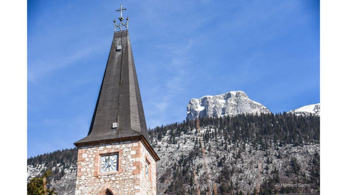 Ein Kirchturm mit einem hohen Dach und einer Uhr steht im Vordergrund. Im Hintergrund sind schneebedeckte Berge und ein klarer blauer Himmel zu sehen. | © Herbert Sams