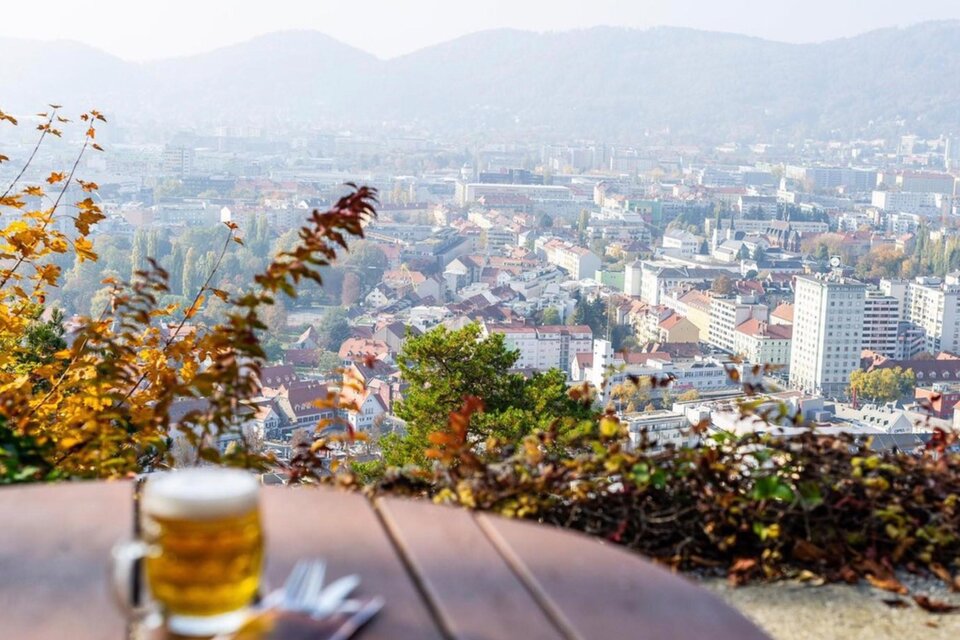 Ein Glas Bier auf einem Holztisch mit Blick auf eine schöne Stadtlandschaft. Im Hintergrund sind Berge und herbstliche Bäume zu sehen. | © -restaurant-schlossberg