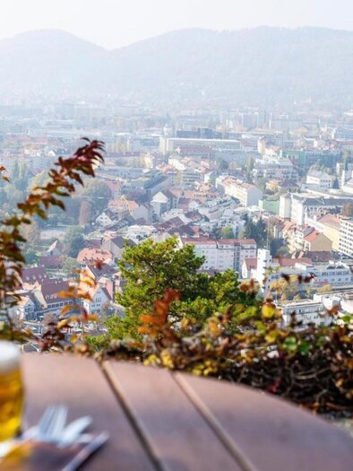 A glass of beer on a wooden table with a view of a beautiful urban landscape. In the background, there are mountains and autumn trees visible. | © -restaurant-schlossberg