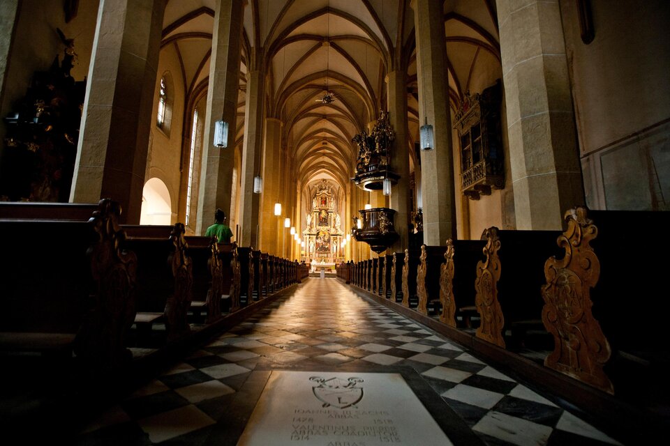 Eine lange Kirche mit hohen gewölbten Decken und Holzbankreihen. Der Boden ist mit einem Schachbrettmuster ausgelegt, das die Perspektive betont. | © TV Murau