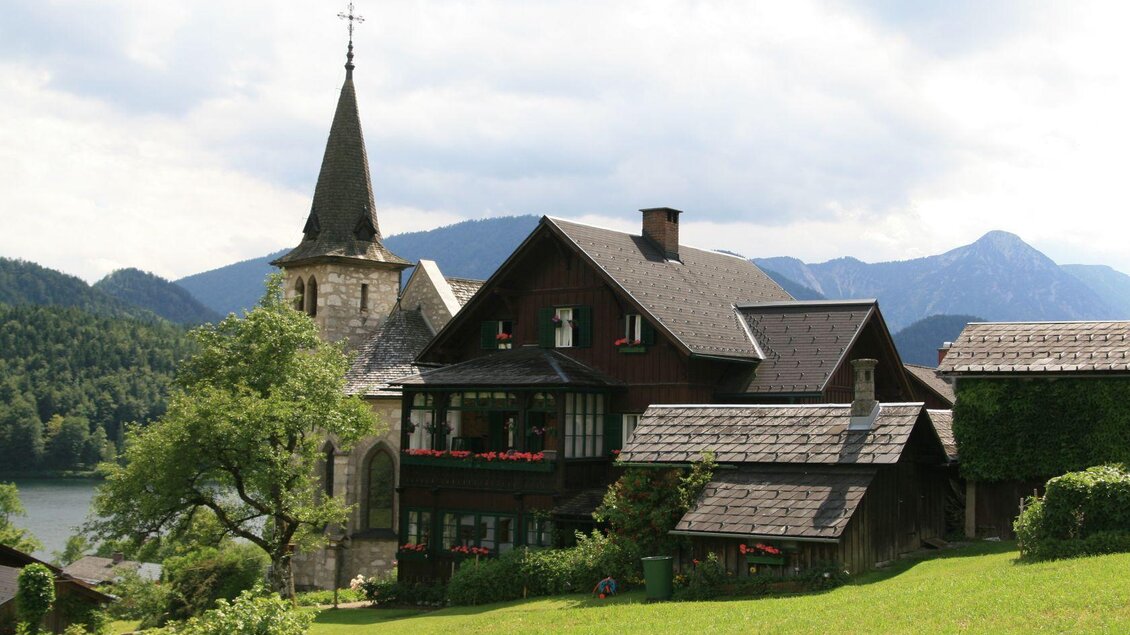 Ein hübsches traditionelles Haus neben einer Kirche, umgeben von grünen Wiesen und Bergen. Der Himmel ist leicht bewölkt, was eine ruhige Stimmung vermittelt. | © Rabensteiner