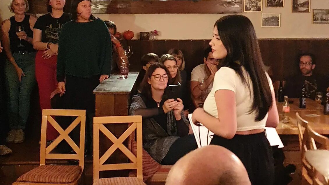 Eine Frau spricht auf einer kleinen Bühne vor Publikum. Im Hintergrund sitzen Zuhörer und schauen interessiert zu. | © Sabine Hermann