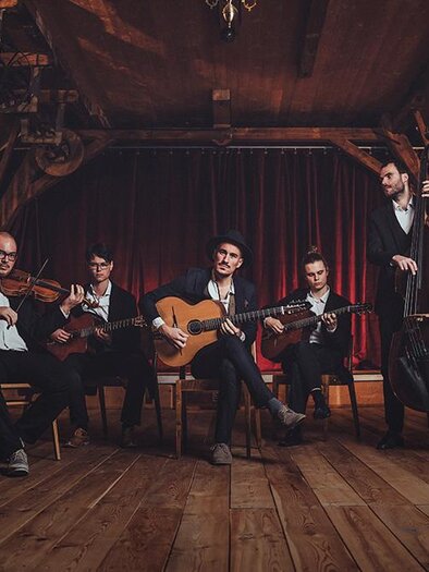 A band performs in a rustic room with a wooden floor. They play various instruments, including guitar, violin, and double bass. | ©  Reithofer Media