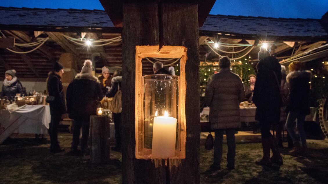 Eine stimmungsvolle Veranstaltung im Freien mit Weihnachtslichtern und Menschen, die sich unterhalten. Im Vordergrund ist eine Laterne mit einer brennenden Kerze zu sehen. | © Klaudia Riebenbauer