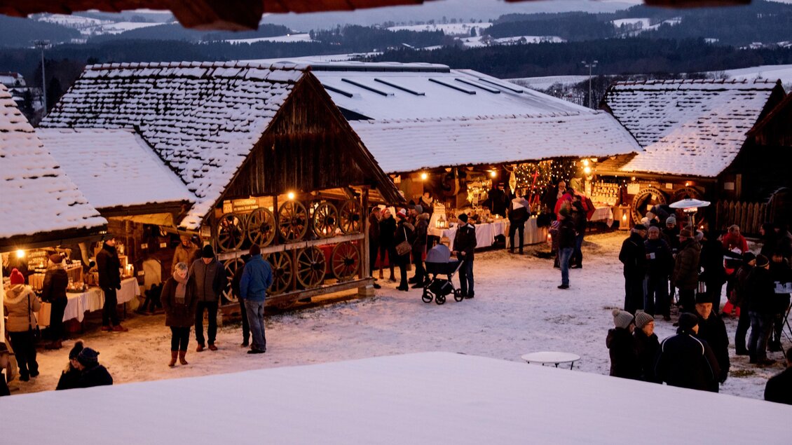 Ein Weihnachtsmarkt mit festlich beleuchteten Ständen in einer verschneiten Umgebung. Besucher schlendern zwischen den Hütten und genießen die frohe Stimmung. | © Klaudia Riebenbauer
