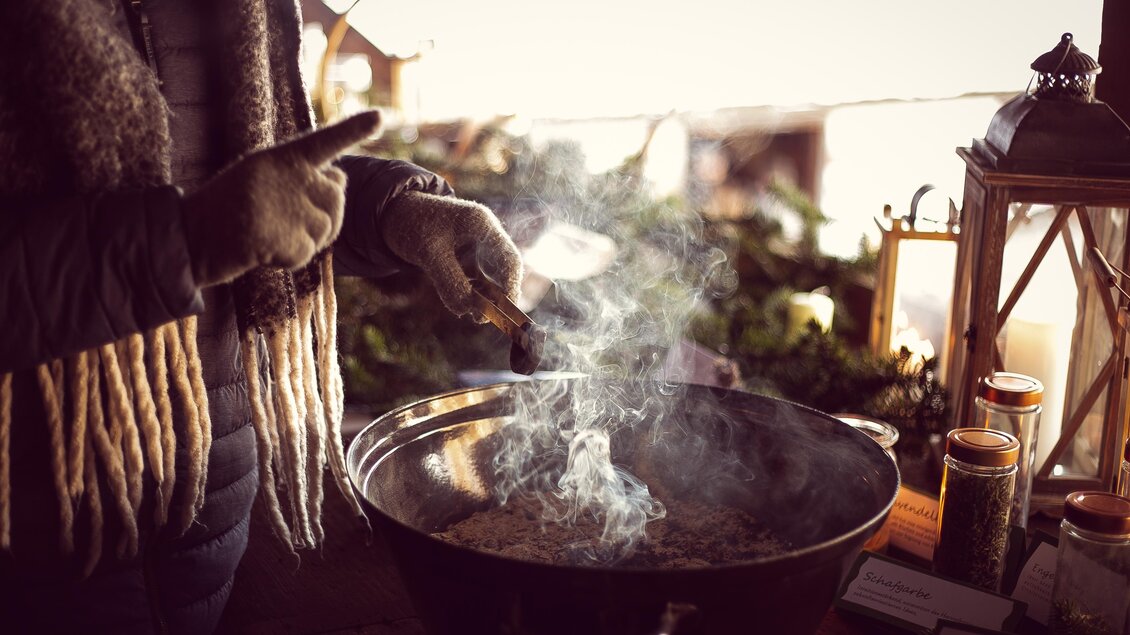 Eine Person rührt in einem großen Topf mit rauchendem Inhalt. Im Hintergrund sind einige dekorative Elemente und Kerzen zu sehen. | © Sonja Spitzer
