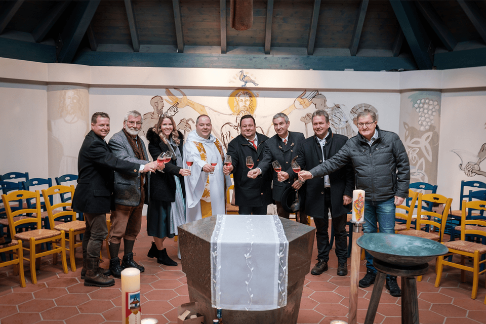 Eine Gruppe von zehn Personen versammelt sich in einem festlichen Raum, um auf ein Ereignis anzustoßen. Im Hintergrund sind Kerzen und eine dekorierte Wand zu sehen. | © Florian Jöbstl