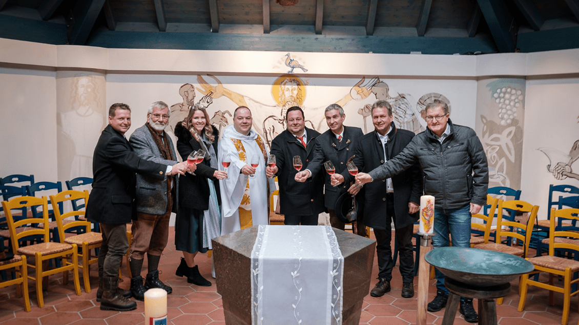 Eine Gruppe von zehn Personen versammelt sich in einem festlichen Raum, um auf ein Ereignis anzustoßen. Im Hintergrund sind Kerzen und eine dekorierte Wand zu sehen. | © Florian Jöbstl