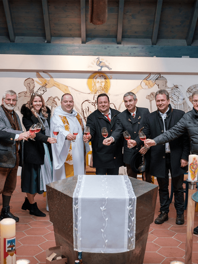 Eine Gruppe von zehn Personen versammelt sich in einem festlichen Raum, um auf ein Ereignis anzustoßen. Im Hintergrund sind Kerzen und eine dekorierte Wand zu sehen. | © Florian Jöbstl