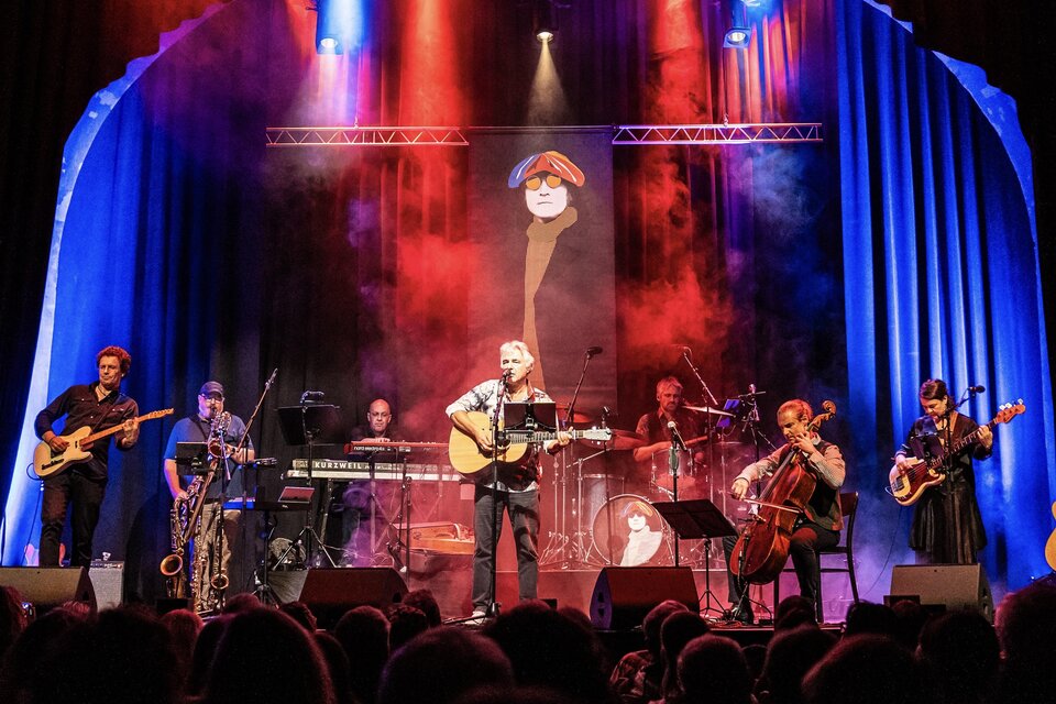 Band auf einer Theaterbühne bei Livekonzert: Sänger in der Mitte mit Akustikgitarre, links und rechts weitere Musiker mit E-Gitarre, Saxophon, Keyboards, Schlagzeug, Cello und Bass; rotes und blaues Bühnenlicht mit Nebel, Publikum als Silhouetten im Vordergrund. | © Georg Cizek-Graf