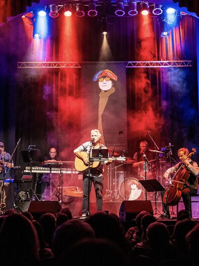 Band auf einer Theaterbühne bei Livekonzert: Sänger in der Mitte mit Akustikgitarre, links und rechts weitere Musiker mit E-Gitarre, Saxophon, Keyboards, Schlagzeug, Cello und Bass; rotes und blaues Bühnenlicht mit Nebel, Publikum als Silhouetten im Vordergrund. | © Georg Cizek-Graf