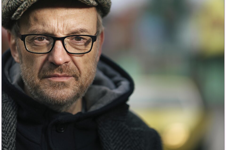 Ein Mann mit Brille und einer Mütze schaut ernst in die Kamera. Im Hintergrund sind verschwommene Farben und Formen zu sehen. | © Josef-Hader-©-www.lukasbeck.com