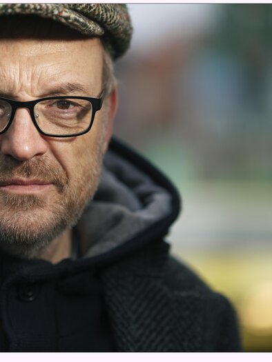 A man with glasses and a hat looks seriously at the camera. In the background, blurred colors and shapes can be seen. | © Josef-Hader-©-www.lukasbeck.com