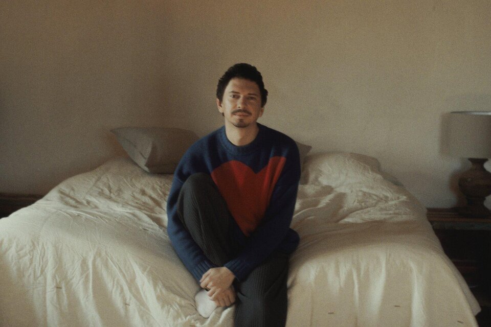 Ein Mann sitzt auf der Bettkante in einem schlicht eingerichteten Schlafzimmer. Er trägt einen blauen Pullover mit rotem Herzen und schaut nachdenklich. | © Julian le Play