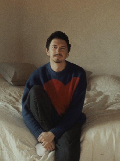 A man is sitting on the edge of the bed in a simply furnished bedroom. He is wearing a blue sweater with a red heart and looks pensive. | © Julian le Play
