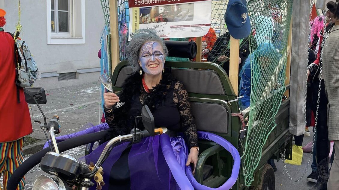 Eine Frau in fantasievoller Verkleidung sitzt in einem Fahrzeug mit einem stilisierten Delfin oben drauf. Um sie herum sind bunte Luftballons und weitere Feiernde zu sehen. | © Stadtgemeinde Köflach