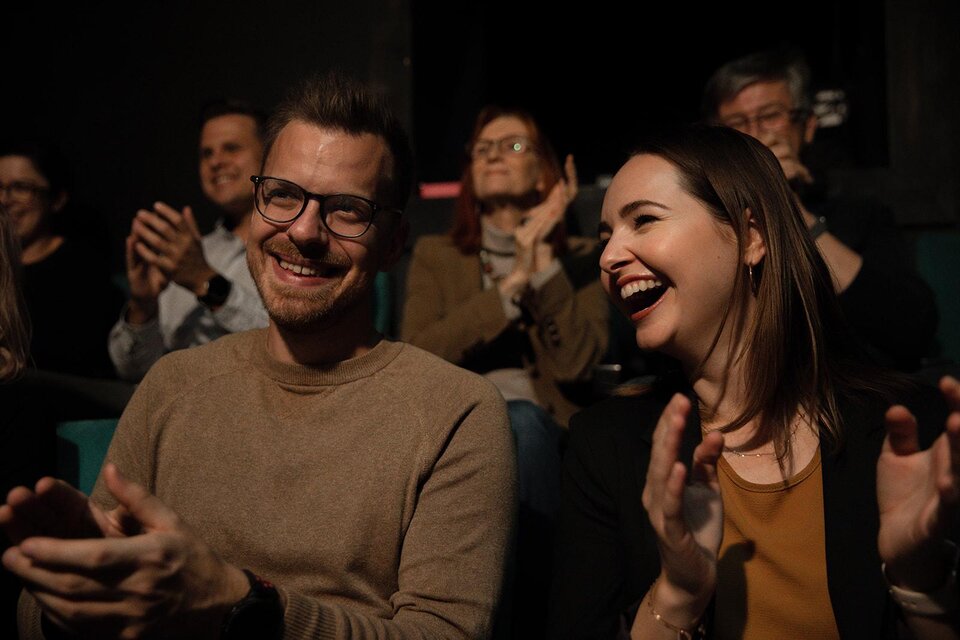 Zwei lachende Menschen im Publikum, die applaudieren. Im Hintergrund sind weitere Zuschauer zu sehen, die ebenfalls Freude ausdrücken.