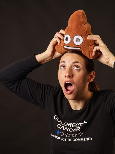 A woman is wearing a stuffed animal shaped like feces on her head and has a surprised facial expression. She is wearing a black T-shirt with the inscription about colon cancer. | © Lukas Fritzer