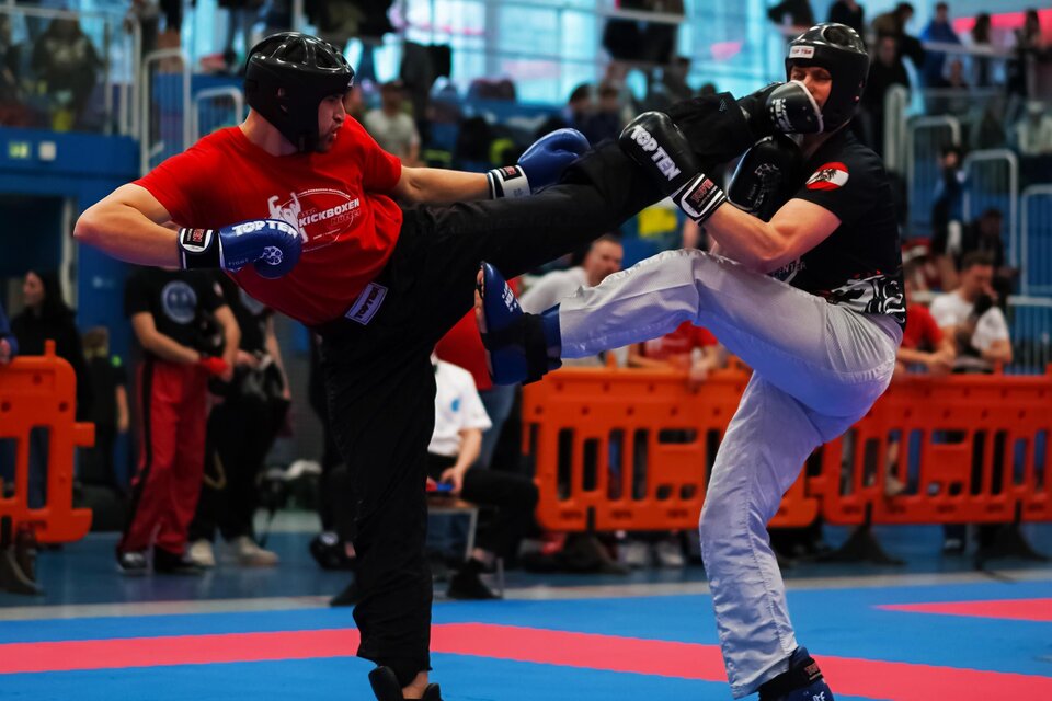 Zwei Kämpfer im Wettkampf, einer in roter und der andere in schwarzer Ausrüstung. Die Atmosphäre ist lebhaft mit Zuschauern im Hintergrund. | © Hütter