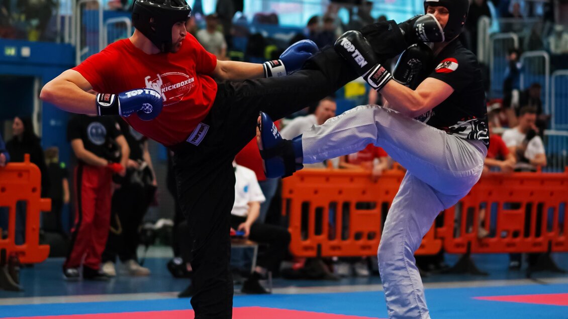 Zwei Kämpfer im Wettkampf, einer in roter und der andere in schwarzer Ausrüstung. Die Atmosphäre ist lebhaft mit Zuschauern im Hintergrund. | © Hütter